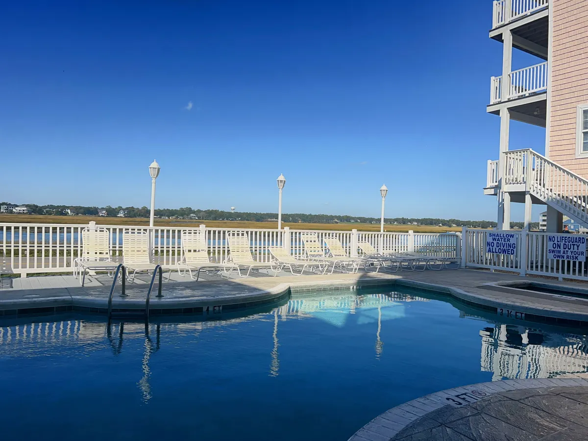 An inviting exterior scene featuring a sparkling swimming pool surrounded by lounge chairs, set against a waterfront view and the villa's building. The area promises relaxation and group socializing in the sun, with open water and blue skies enhancing the leisure atmosphere.