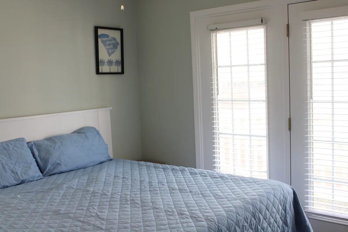 A tranquil bedroom scene with a bed dressed in a blue quilt, a window with blinds allowing sunlight to filter in, and a peaceful ambiance. The composition invites the viewer to imagine enjoying a quiet morning with coffee while soaking in views from the comfort of their room.