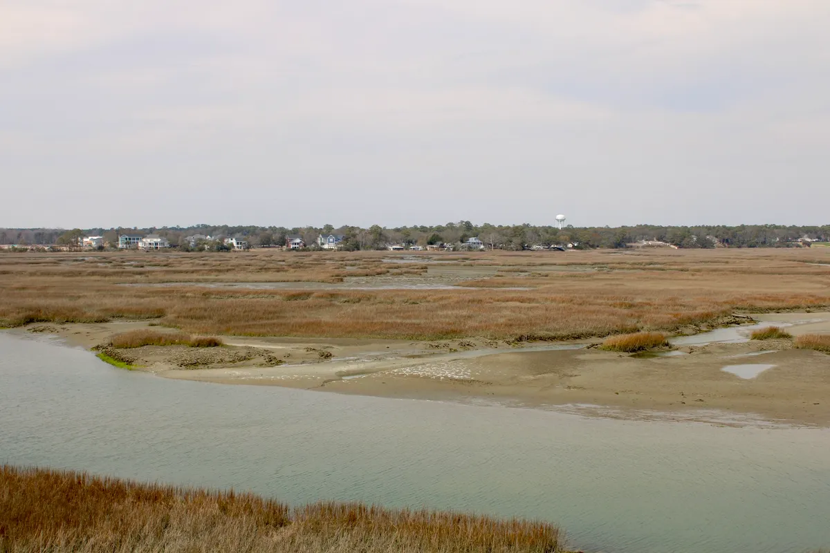 Expansive marshland and water under a wide, open sky, with distant houses dotting the horizon. The natural landscape invites outdoor activities and exploration, evoking a sense of coastal adventure and relaxation.