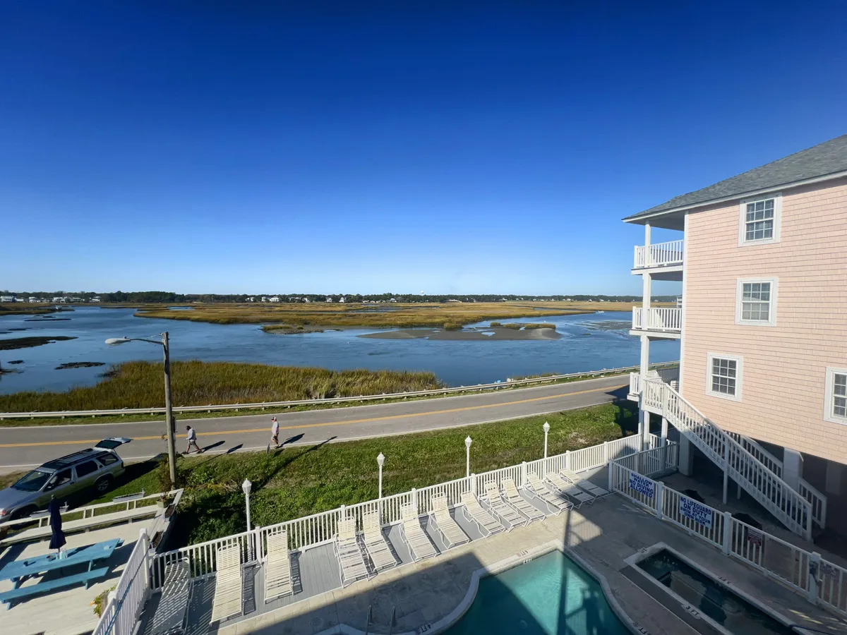 A panoramic view from the property encompasses the waterfront, swimming pool, outdoor seating, and a road with pedestrians. The vista showcases the proximity to the water and the lively yet tranquil environment, ideal for sunsets and social gatherings.