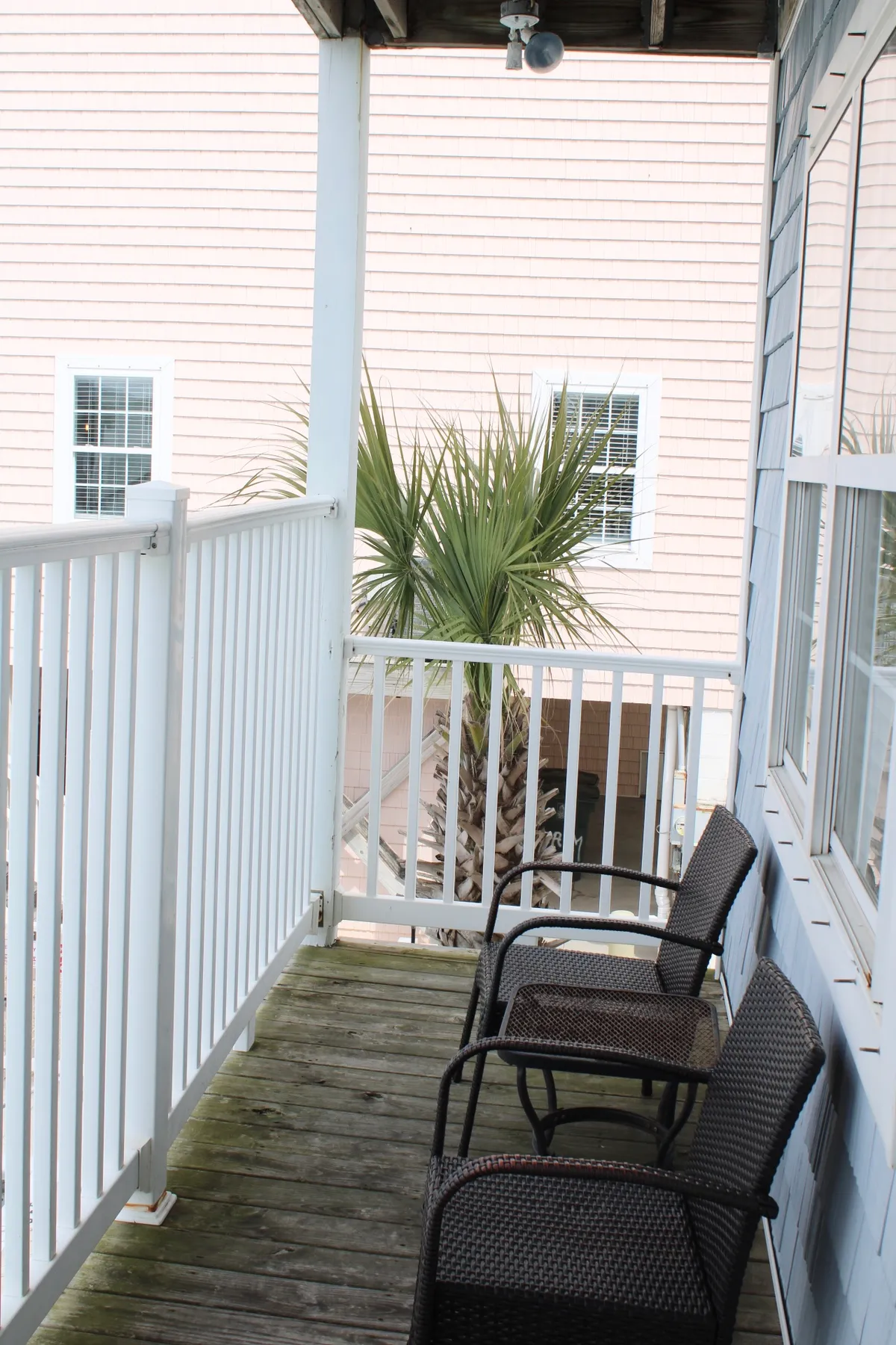 An exterior view featuring a balcony deck furnished with two wicker chairs, shaded by a palm tree. The wooden deck provides a tranquil spot to enjoy the sunrise or unwind under the stars, echoing the villa's focus on outdoor living.