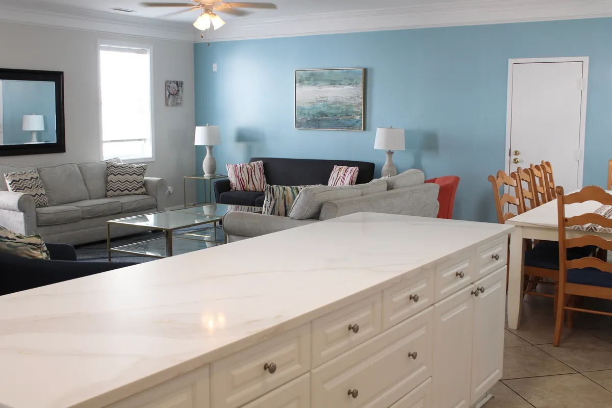 This image captures the open-plan living area, featuring comfortable sofas, a central coffee table, a dining table, kitchen island, and decorative artwork. The space is brightly lit and designed for both relaxation and socializing, with the kitchen and dining area seamlessly integrated into the living room.