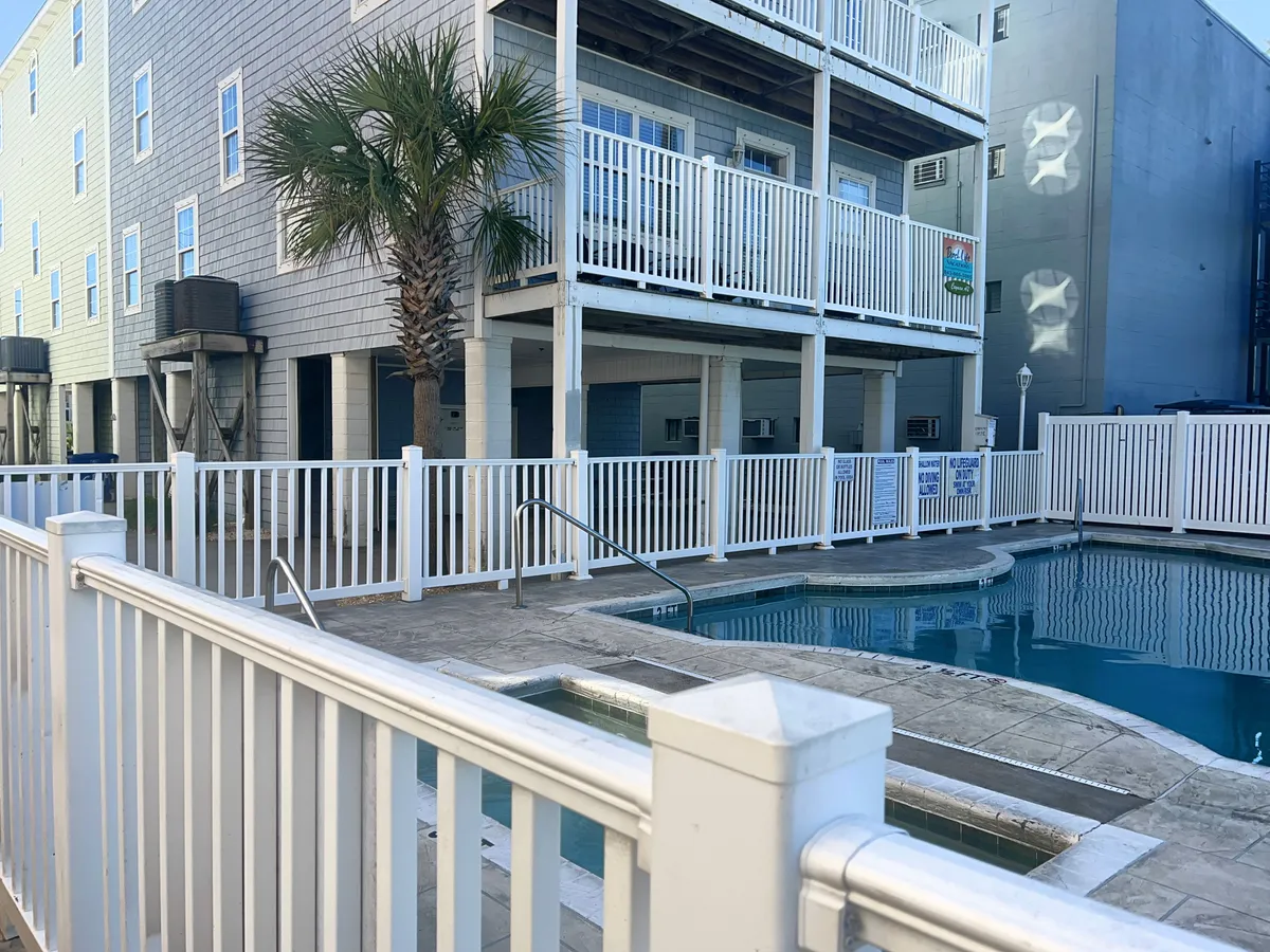 This exterior image showcases a sparkling swimming pool surrounded by lounge chairs, a palm tree, and a balcony with fencing. The pool area is bright and inviting, positioned to catch the coastal sun, and hints at the leisurely atmosphere of the property. The presence of palm trees and open sky evokes a classic beach vacation vibe.