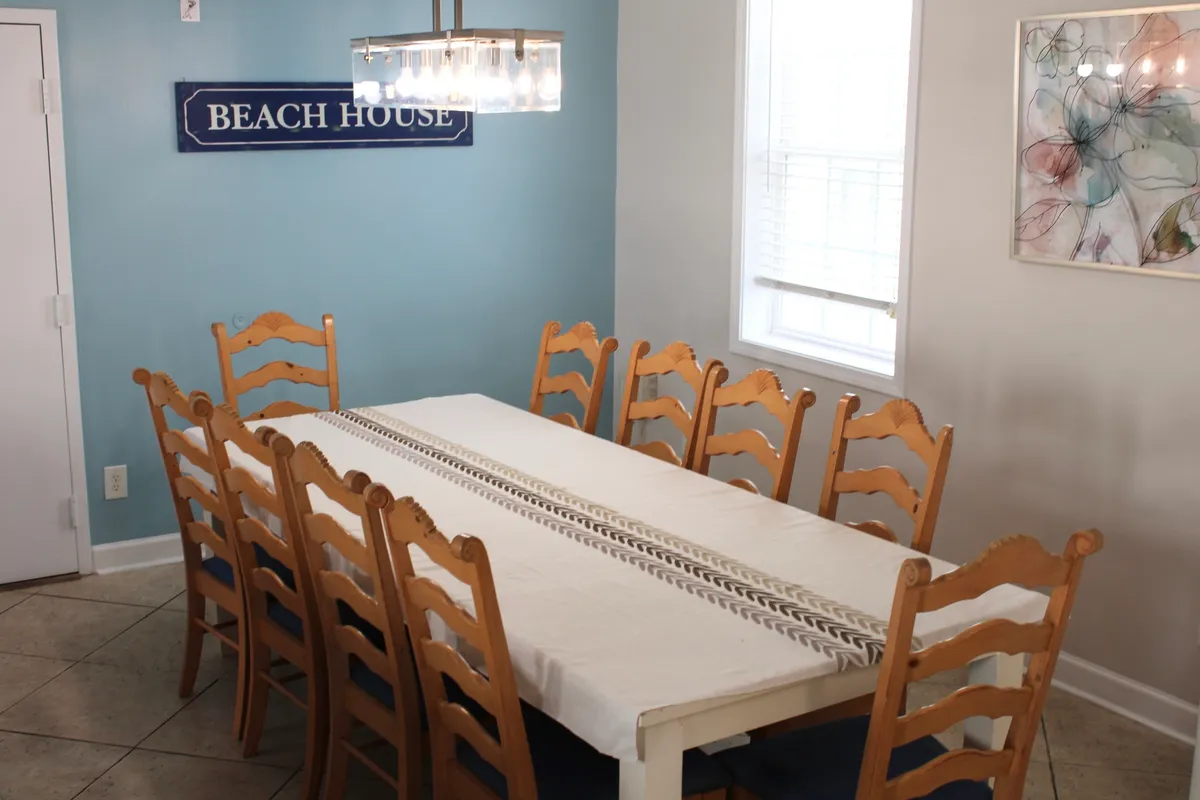 A long dining table sits under a striking chandelier, with space for a large group. The BEACH HOUSE sign and decorative artwork create a laid-back yet celebratory atmosphere, perfect for family meals, celebrations, and festive gatherings.