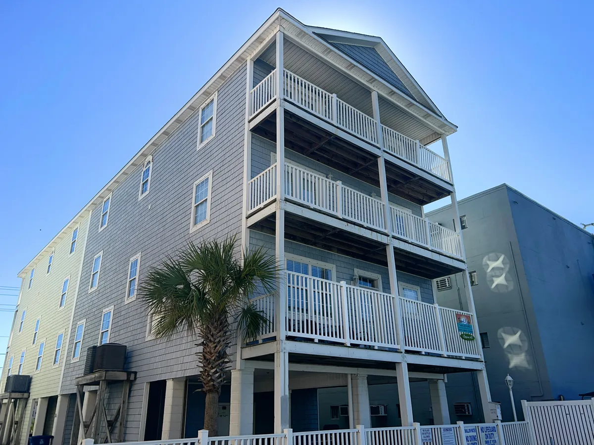 The hero image showcases the grand exterior of Grand Cayman Villa A2, featuring multiple spacious balconies with white railings, lush palm trees, inviting light blue siding, and a welcoming front porch. The scene captures the cheerful, relaxed atmosphere of a beachside retreat, with abundant outdoor spaces ideal for gatherings and unwinding.