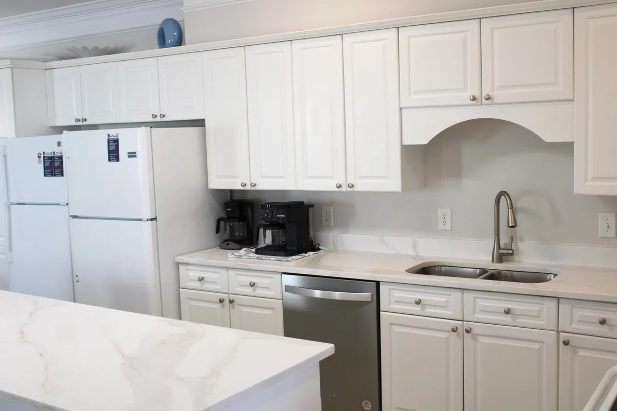 A spacious and inviting gourmet kitchen featuring sleek white cabinetry, stainless steel appliances, a marble countertop, and a coffee maker. The setting is perfect for group meal preparation, socializing, and culinary experiences together.