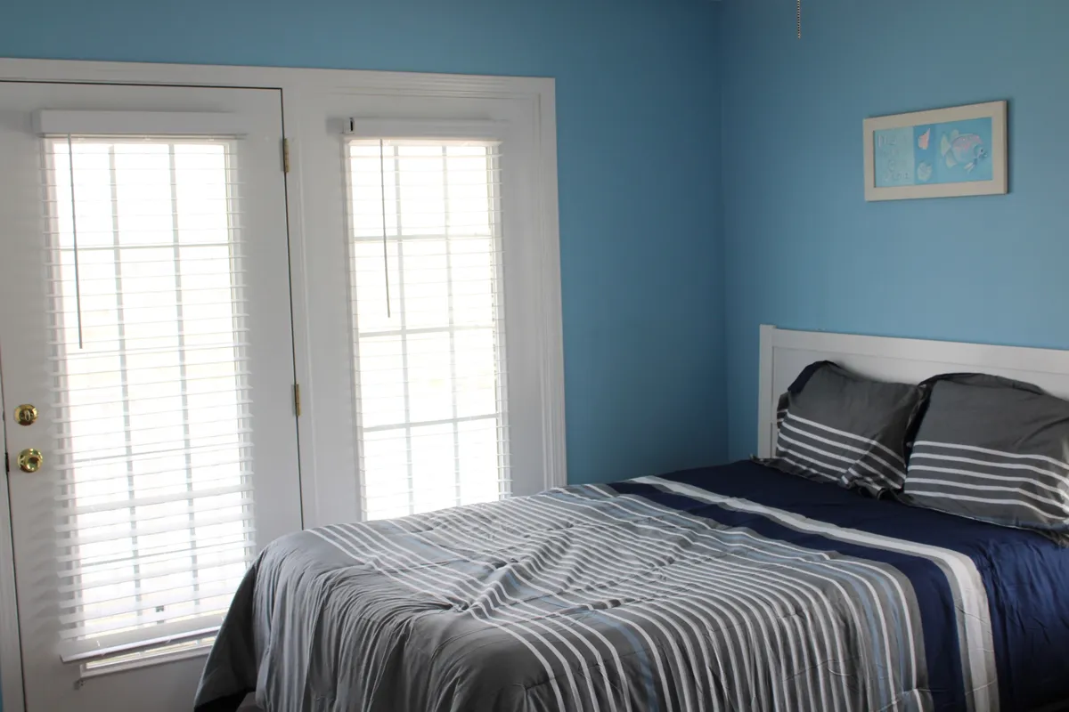 This image features a serene bedroom with striped bedding and double doors that open directly outside. Morning light streams into the room, suggesting easy indoor-outdoor flow and creating a sense of connection to the surrounding environment.