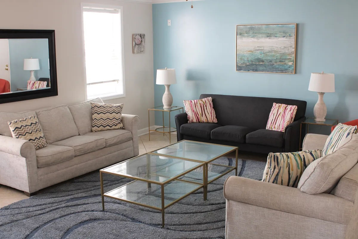A welcoming living area featuring a glass coffee table, sofas adorned with decorative pillows, framed artwork, and abundant morning sunlight streaming through the windows. The space exudes an inviting and social atmosphere ideal for group relaxation or planning outdoor excursions.