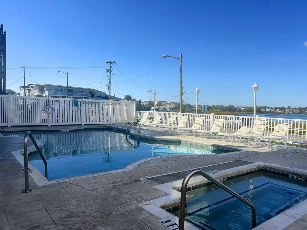 An inviting exterior scene featuring a swimming pool, hot tub, lounge chairs, a secure fence, and a lush surrounding landscape. The area is ideal for sunbathing, socializing, and unwinding after beach adventures, with ample seating to accommodate groups or families.