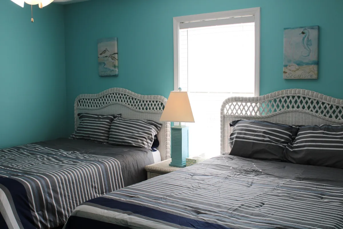 This image features two beds with striped bedding set against a window that lets in abundant natural light. A decorative lamp and a touch of simple artwork complete the serene and welcoming atmosphere, perfect for a restful night or a peaceful morning.